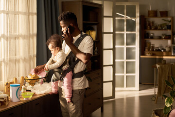Black young adult man multitasking holding infant child in baby carrier, while talking on smartphone and preparing baby food in home kitchen during paternity leave morning routine
