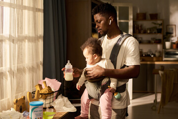Black young adult man holding infant in baby carrier preparing baby bottle in kitchen during daytime paternity leave concept, showing father caring for child at home multitasking