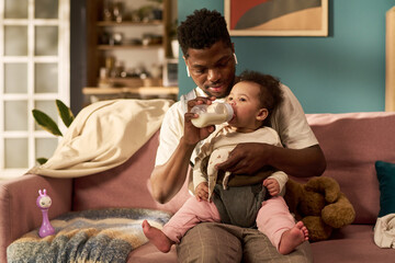 Black young adult man feeding infant with bottle while sitting on sofa, holding baby in lap, demonstrating paternity leave and nurturing fatherhood in home setting