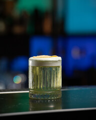 refreshing yellow-green alcoholic cocktail, topped with a white foam and a dehydrated apple slice, is served in a textured glass on a dark green bar counte