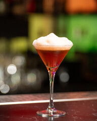 A deep amber or brown alcoholic cocktail, topped with a thick layer of white foam, is presented in...