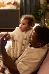 Black man holding child on lap, smiling and interacting together in home setting, demonstrating...
