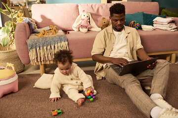 Black young adult man sitting on floor using laptop while caring for infant child playing with toys...