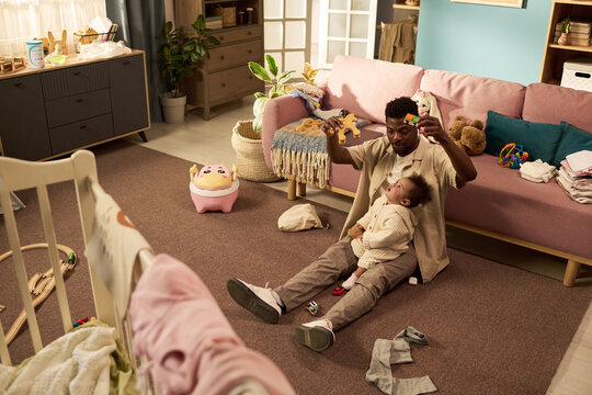 Black young adult man sitting on floor holding infant child in arms playing with soft toy in living room, surrounded by baby items representing paternity leave concept