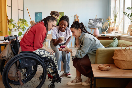 Three young adult women, including Black and Caucasian, gathering in art studio, smiling and interacting with smartphone, one woman in wheelchair with disability participating in conversation