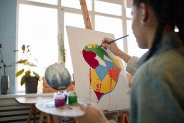 Young woman painting colorful abstract heart on canvas using paintbrush, holding palette in hand,...