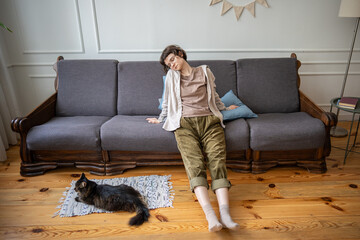 Exhausted indifferent woman lounging on sofa with cat resting nearby on floor in lethargy and...