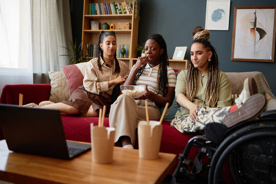 Three young adult women, including Black and Caucasian, sitting on sofa watching laptop screen together, one woman with disability using wheelchair, sharing popcorn and drinks