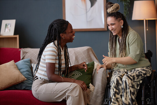 Young adult Black woman sitting on sofa laughing with young adult Caucasian woman with disability in wheelchair, both smiling and gesturing while enjoying friendly conversation indoors - Powered by Adobe