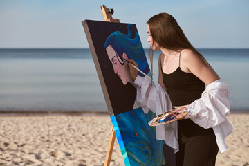 Young female artist stands on tranquil beach, intently painting portrait. Calm sea and open sky set...