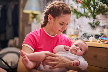 Young woman with braided hair holds infant in cozy room. Warm light casts gentle glow. Christmas...