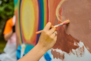 Young caucasian female uses paintbrush to create vibrant mural on outdoor wall. Scene captures...