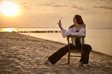 Young woman, styled as painter, sits contemplatively on chair at beach during sunset. Serene...