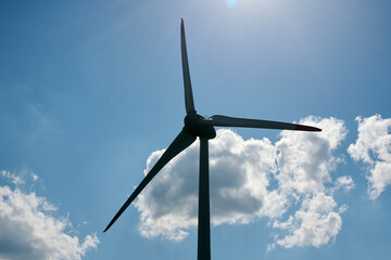 Silhouetted wind turbine set against clear blue sky with soft white clouds scattered across...