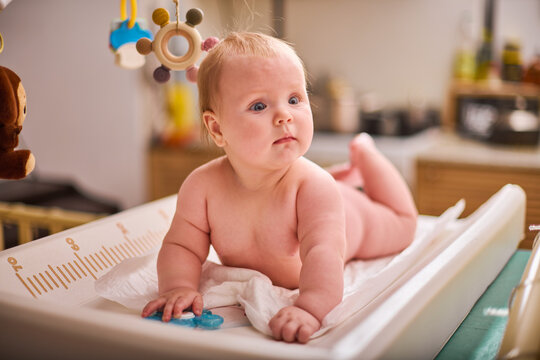 Baby girl with short blonde hair, lying on changing table surrounded by toys. Soft warm lighting enhances her curious expression in inviting nursery