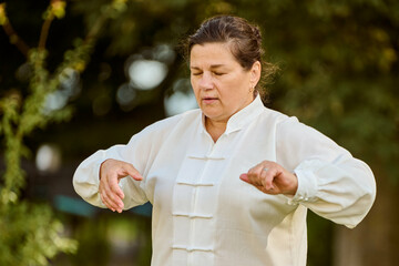 Mature caucasian woman with brown hair performs qigong. Her eyes closed, expression serene, wearing...