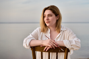 Young woman in white blouse sits on chair by serene beach. Sunset casts warm glow, evoking calm and introspection. Setting highlights beauty of nature and peaceful solitude