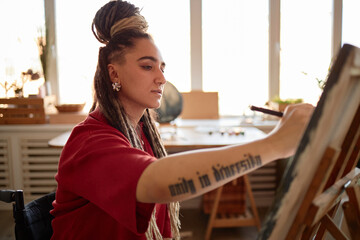 Young adult Caucasian woman with disability painting on canvas in bright studio, showing tattooed...