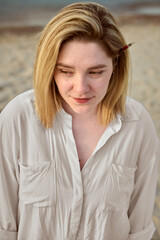Young woman with blonde hair stands pensively on serene beach. Her relaxed expression reflects peaceful, introspective mood. Soft, sandy background suggests tranquility