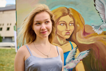 Young woman with blonde hair and light skin standing near vibrant artwork. Outdoor setting with bright lighting. Art piece features serene woman and bird