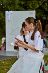 Young adult caucasian women painting outdoors at festival. Intense focus, creative atmosphere, brushes in hand. Background features greenery and easel
