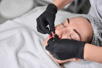 Young adult woman receives lip treatment in tranquil spa setting. Black-gloved hands expertly apply precision techniques, enhancing relaxation and self-care. Ambiance is soothing and meticulous