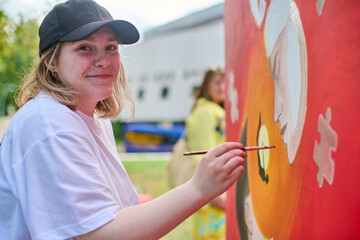 Young caucasian woman in black cap painting vibrant mural outdoors. Bright daylight and green surroundings enhance focus, with modern building in backdrop