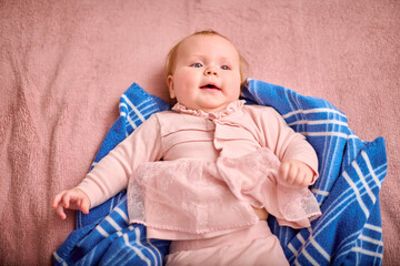 Baby girl, child, blonde hair, caucasian, wearing pink dress, lying on soft, blue-checkered blanket. Cozy environment, warm lighting, relaxed expression, indoor setting