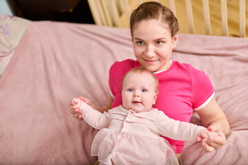 Young adult female with brown hair holding adorable baby girl in pink outfit. European ethnicity, both smiling indoors on soft pink blanket with wooden crib nearby