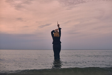 Woman stands waist-deep in tranquil ocean at sunset, raising her arms in contemplation. Peaceful setting, with pastel skies, evokes sense of freedom and introspection
