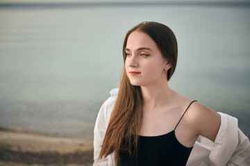 Young woman stands contemplatively by serene seaside, with gentle waves in background. Her relaxed expression and flowing hair capture peaceful, introspective mood