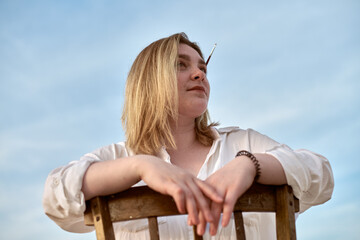 Young woman gazes thoughtfully under clear sky, leaning on wooden chair. With paintbrush in her hair, she embodies creativity and calm, suggesting introspection and inspiration