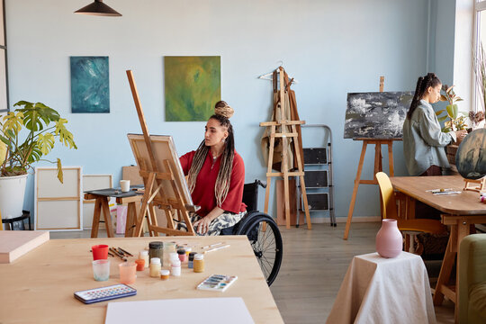 Young adult Caucasian woman with disability painting at easel in art studio while young woman standing in background working on canvas, creative workspace with art supplies visible