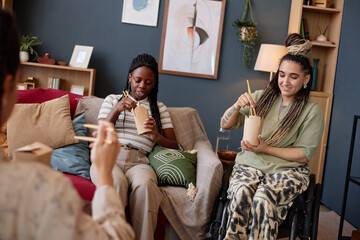 Young adult Black woman and Caucasian woman with disability sitting on sofa and wheelchair eating takeout food with chopsticks, talking with unseen friend at home