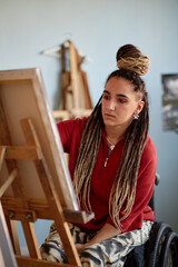 Young adult Caucasian woman with disability sitting in wheelchair painting on canvas, focused...