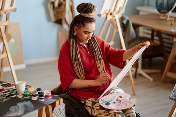 Young adult Caucasian woman with disability sitting in wheelchair painting on canvas using...