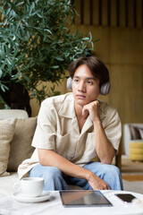 Non binary Asian student man sitting in modern coffee shop wearing headphones
