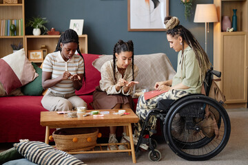 Three young adult women, one Caucasian with disability in wheelchair, sitting together creating...