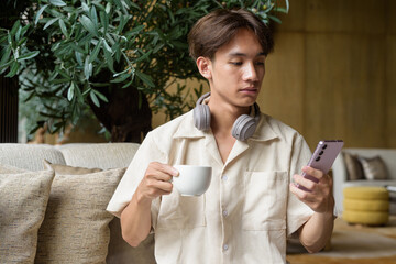 Non binary Asian student man sitting in modern cafe using phone and drinking coffee