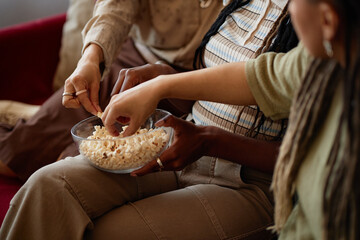 Group of young women sharing popcorn, sitting close together, reaching into glass bowl with hands,...