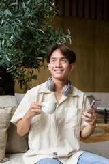 Non binary Asian student man sitting in modern cafe using phone and drinking coffee