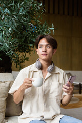 Non binary Asian student man sitting in modern cafe using phone and drinking coffee