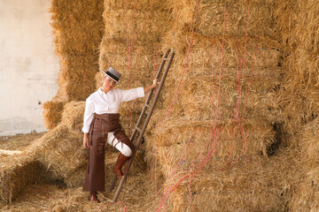 Young, pretty, blonde woman in white shirt, leather chaps, riding boots and hat, holding on to a ladder where she keeps the hay bales for the horses inside the stable. Concept animals, riding, horses.