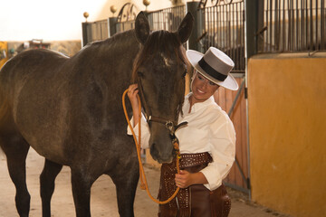 Young, beautiful, blonde woman in white shirt, leather chaps and hat, posing looking at her horse, inside the stable. Concept animals, riding, horses, sport.