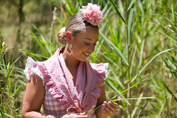 Portrait of young woman, beautiful, blonde, with typical Andalusian flamenco costume to go on a pilgrimage with small shawl and pink flower, posing in the middle of the field.