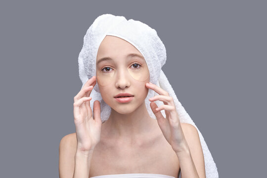 Young caucasian female with under eye patches and towel on head, gray background, skincare routine. - Powered by Adobe
