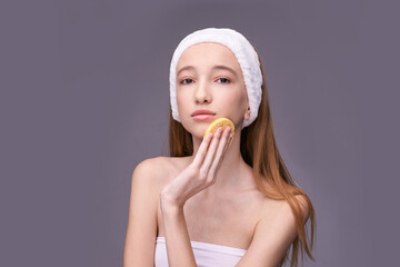 Young caucasian female skincare routine with headband and sponge against gray background.