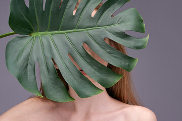 Mysterious young caucasian woman partially hidden behind large monstera leaf against gray background.