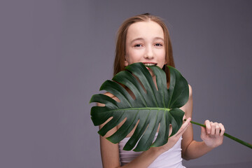 Young caucasian female teen with long hair holding large monstera leaf against gray background and smiling.