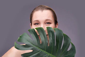 Smiling caucasian adult female peeking from monstera leaf against gray background.